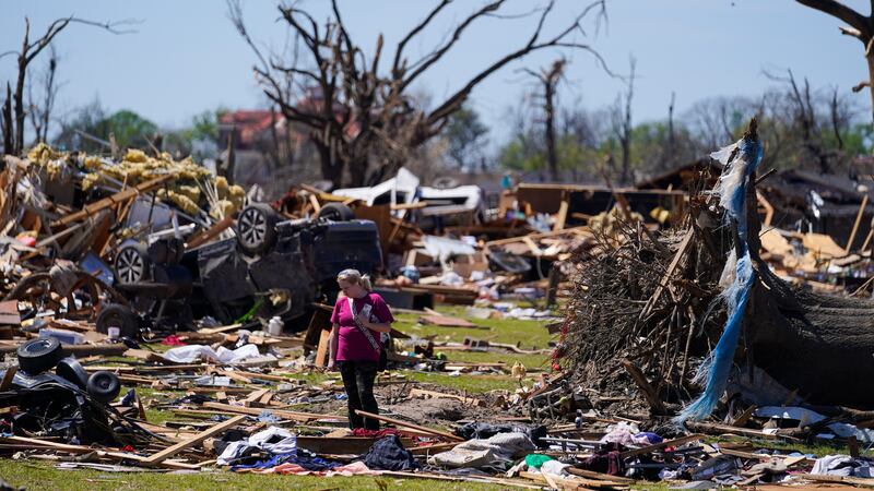 A woman walks near an uprooted tree, a flipped vehicle and debris from homes damaged by a...